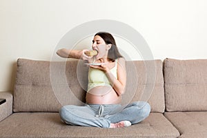 Excited pregnant woman is eating a slice of cake resting on the sofa at home. Love to sweet during pregnancy