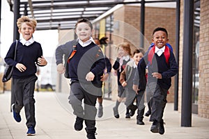 Excited primary school kids, wearing school uniforms and backpacks, running on a walkway outside their school building, front view