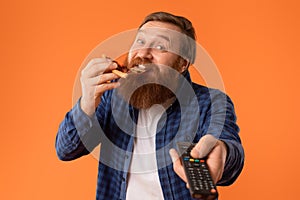 Excited redhaired bearded man enjoys pizza watching TV, orange background