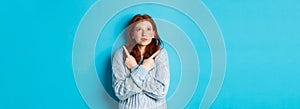 Excited redhead girl pointing fingers sideways, showing two choices and looking tempted up, standing against blue