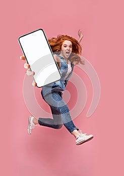 Excited redhead woman having fun, jumping,demonstrating mobile phone with empty white screen on pink background