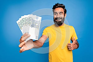 Excited rich indian middle aged man holding euro money and gesturing thumb up, celebrating big financial luck