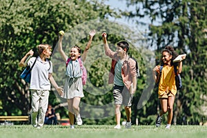 Excited schoolkids gesturing with raised hands while running on lawn in park