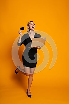 Excited screaming woman holding laptop and credit card isolated over orange background