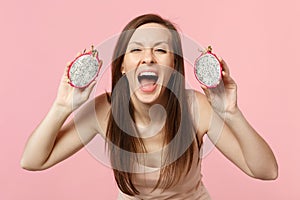 Excited screaming young woman holding halfs of fresh ripe pitahaya, dragon fruit isolated on pink pastel wall background