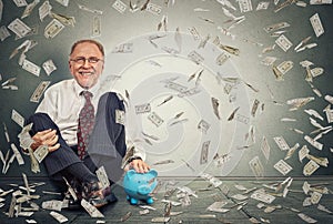 Excited senior man sitting on a floor with piggy bank under a money rain