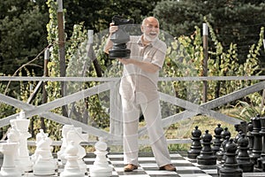 Excited senior playing chess on big chessboard in the park. Chess game for old man. Older grandfather, grandpa pensioner