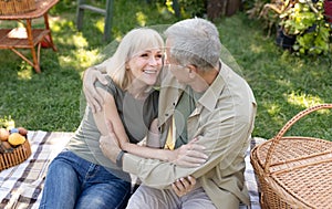 Excited senior spouses having picnic in garden, embracing and laughing, sitting together on blanket outdoors