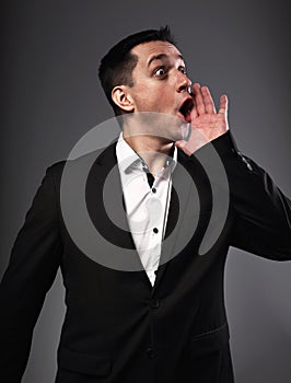 Excited shouting young business man gesturing the hands loudspeaker sign in celebrating emotion with wide open mouth to agitate on