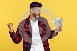 Excited smiling bearded hipster young man in plaid shirt holding money cash and celebrate over yellow background.