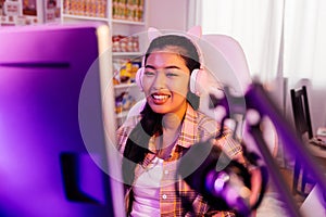 Excited and smiling gamer girl in cute headset with mic playing an online video game. Young Asian woman talking to