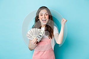 Excited smiling girl fist pump and hold money prize, winning cash, receive income from something, standing happy against