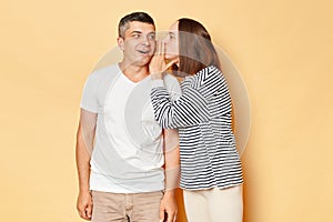 Excited smiling man hearing his wife whispering saying something to ear wearing casual attires standing isolated over beige