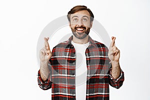 Excited smiling man looking up with hope, making wish, cross fingers for good luck, standing over white background