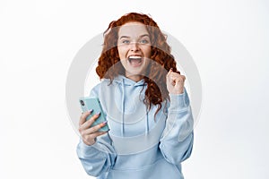 Excited smiling redhead girl rejoicing of winning, making money on mobile phone, holding smartphone and making fist pump