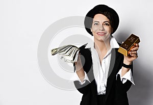 Excited smiling woman in stylish hat and black jacket demonstrates a fan of dollars and a bar of gold in her hands