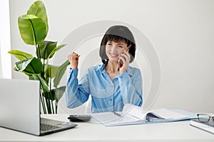 Excited successful employee businesswoman talking on smartphone say yes doing winner gesture, clench fist, sit at office