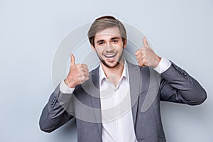 Excited successful handsome young man in formal wear showing gesture like with beaming smile while standing on gray