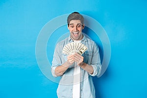 Excited successful man counting money, looking satisfied at cash and smiling, standing over blue background