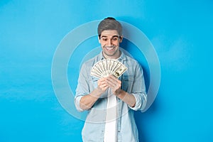 Excited successful man counting money, looking satisfied at cash and smiling, standing over blue background