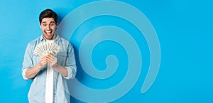 Excited successful man counting money, looking satisfied at cash and smiling, standing over blue background