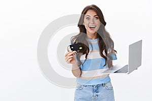 Excited surprised and amused attractive female, smiling impressed, hold laptop and black credit camera astonished how