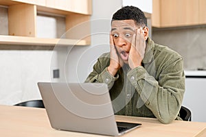 Excited surprised latin dark-haired man in casual shirt staring at laptop. Shocked happy multiracial male entrepreneur