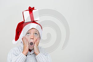 Excited surprised little boy in red santa hat with gift box in head looking upwards. Isolated on white background, banner copy