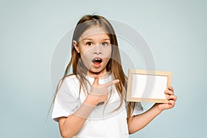 Excited surprised little girl pointing to an empty space in a frame on a blue background. Copy space. Place for