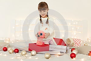 Excited surprised little girl wearing white sweater holding pink soft toy in hands, looking at her gift with big eyes, sitting on