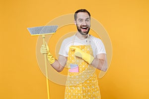 Excited surprised young man househusband in apron rubber gloves doing housework isolated on yellow wall background