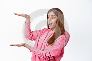 Excited teen girl showing big empty space, holding box copy space, looking at copyspace, standing over white background