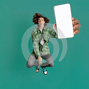 Excited teen guy in casual wear jumping and showing cellphone with empty screen on turquoise studio background, mockup