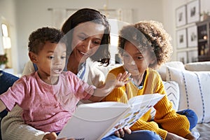 Excited toddler boy sitting on his motherï¿½s knee reading a book in the living room with his older sister, close up