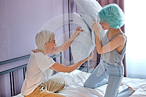 Excited two caucasian women lovely couple sitting on white bed Playing with pillows