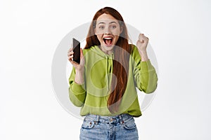 Excited winning ginger girl celebrating, winning money on mobile phone, holding smartphone and triumphing, scream yes