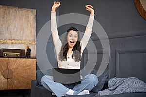 Excited woman celebrating success, raising hands up, sitting on sofa with laptop and reading great news, screaming yes