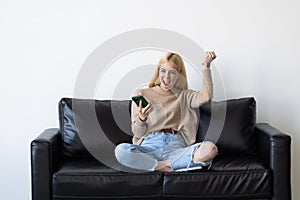 Excited woman checking phone online content celebrating good news sitting on a couch in the living room at home