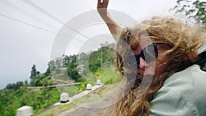 Excited woman leans out car window, hair blowing in wind on mountain road. Female traveler feels freedom, adventure