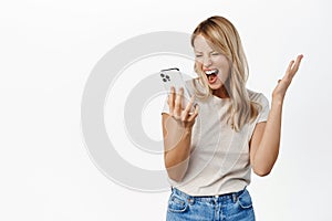 Excited woman looking at mobile phone screen and shouting, screaming at smartphone display, standing over white