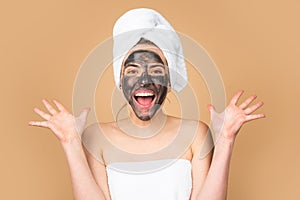 Excited woman with naked shoulder and clay mask on face, on blue. Beautiful model applying cosmetic cream treatment on