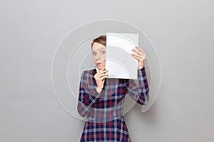 Excited woman is peeking out from behind paper sheet with widened eyes