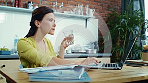 Excited woman reading good news on laptop. Girl drinking water in kitchen.