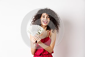 Excited woman in red dress winning money, showing dollar bills and smiling happy, standing on white background