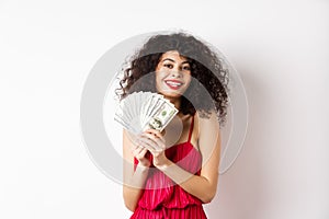 Excited woman in red dress winning money, showing dollar bills and smiling happy, standing on white background
