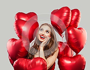 Excited woman with red heart balloons on white background. Happy surprised model girl with red lips makeup, long curly hair. Gift