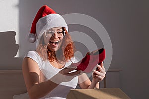 An excited woman in a Santa Claus hat takes red shoes from a cardboard box. The girl unpacks the delivered order for