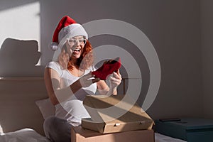 An excited woman in a Santa Claus hat takes red shoes from a cardboard box. The girl unpacks the delivered order for