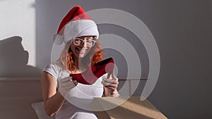 An excited woman in a Santa Claus hat takes red shoes from a cardboard box. The girl unpacks the delivered order for