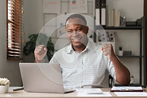 Excited young african american business man working with laptop computer while triumphing with raised hands because